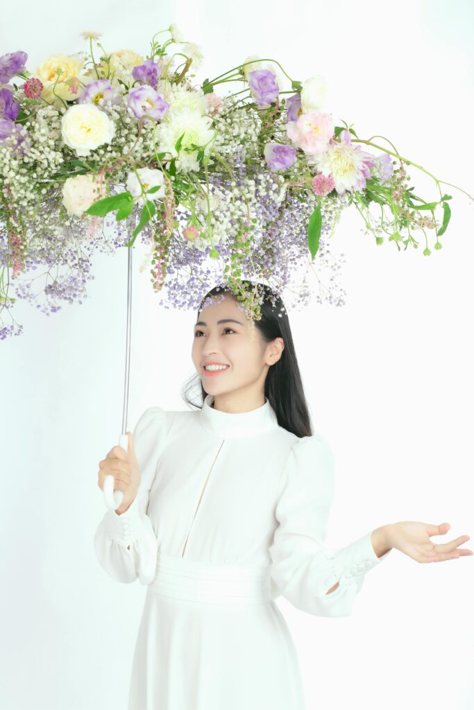 A woman in white smiles while holding a creative umbrella adorned with flowers.